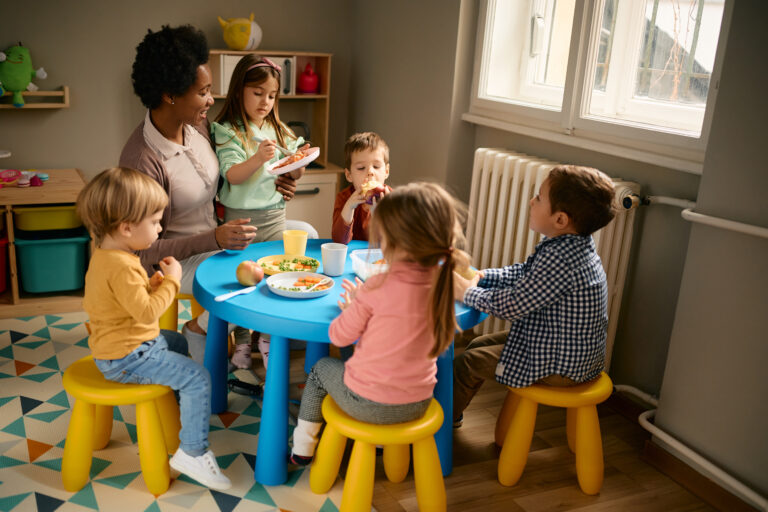 Happy African American female teacher and group of preschool kids having lunch together.