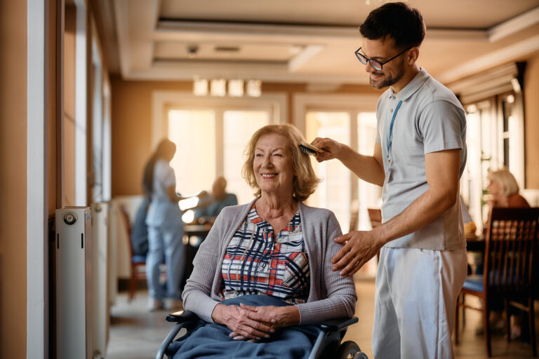 Happy senior woman in wheelchair enjoying while male nurse is brushing her hair at residential care home.
