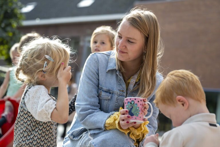 A young woman smiling at a child while other children play around on a sunny day.